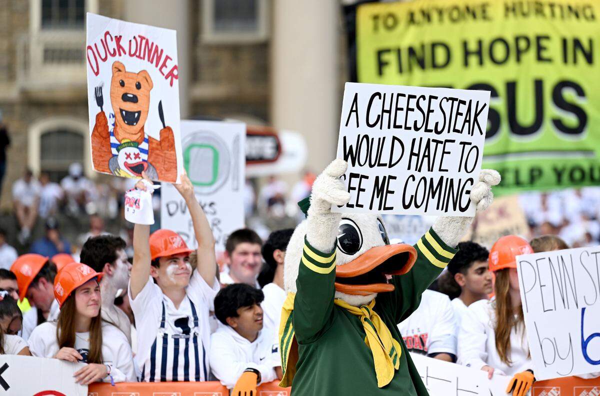 A fan holds up a sign that says “Duck Dinner” as the Oregon duck tries to block him from the cameras during ESPN’s College GameDay at Penn State on Saturday, Sept. 27, 2025.  