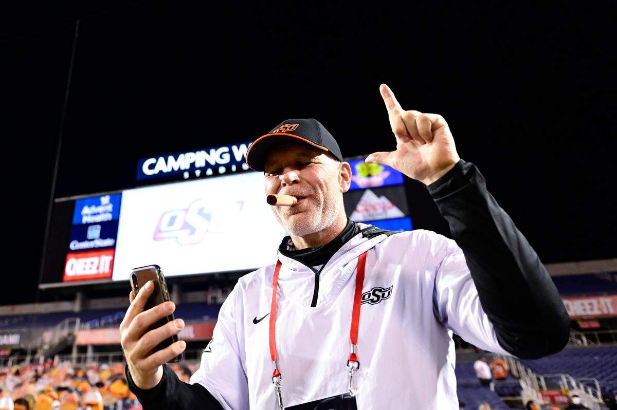 Dec 29, 2020; Orlando, FL, USA; Oklahoma State Cowboys defensive coordinator Jim Knowles (center) enjoys a cigar and talks on the phone after defeating the Miami Hurricanes to win the Cheez-It Bowl Game at Camping World Stadium. Mandatory Credit: Douglas DeFelice-USA TODAY Sports