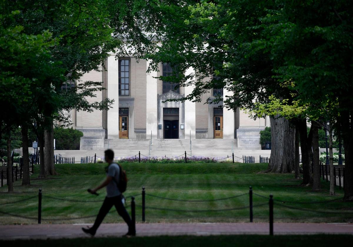 A man wearing a mask walks along Pollock Road with Pattee Library in the distance on the Penn State campus on Thursday, July 16, 2020.