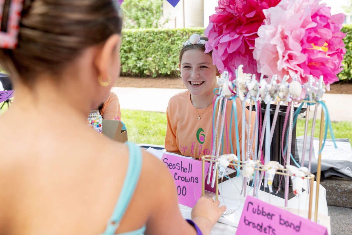 Ryleigh Fortney, 15, chats with a customer as she looks at seashell crowns during the Children and Youth Day sidewalk sale on Wednesday, July 8, 2026. Fortney and her friend Maddie Hatch were selling an assortment of seashell crowns and wands along with tissue paper flowers and beaded bracelets.