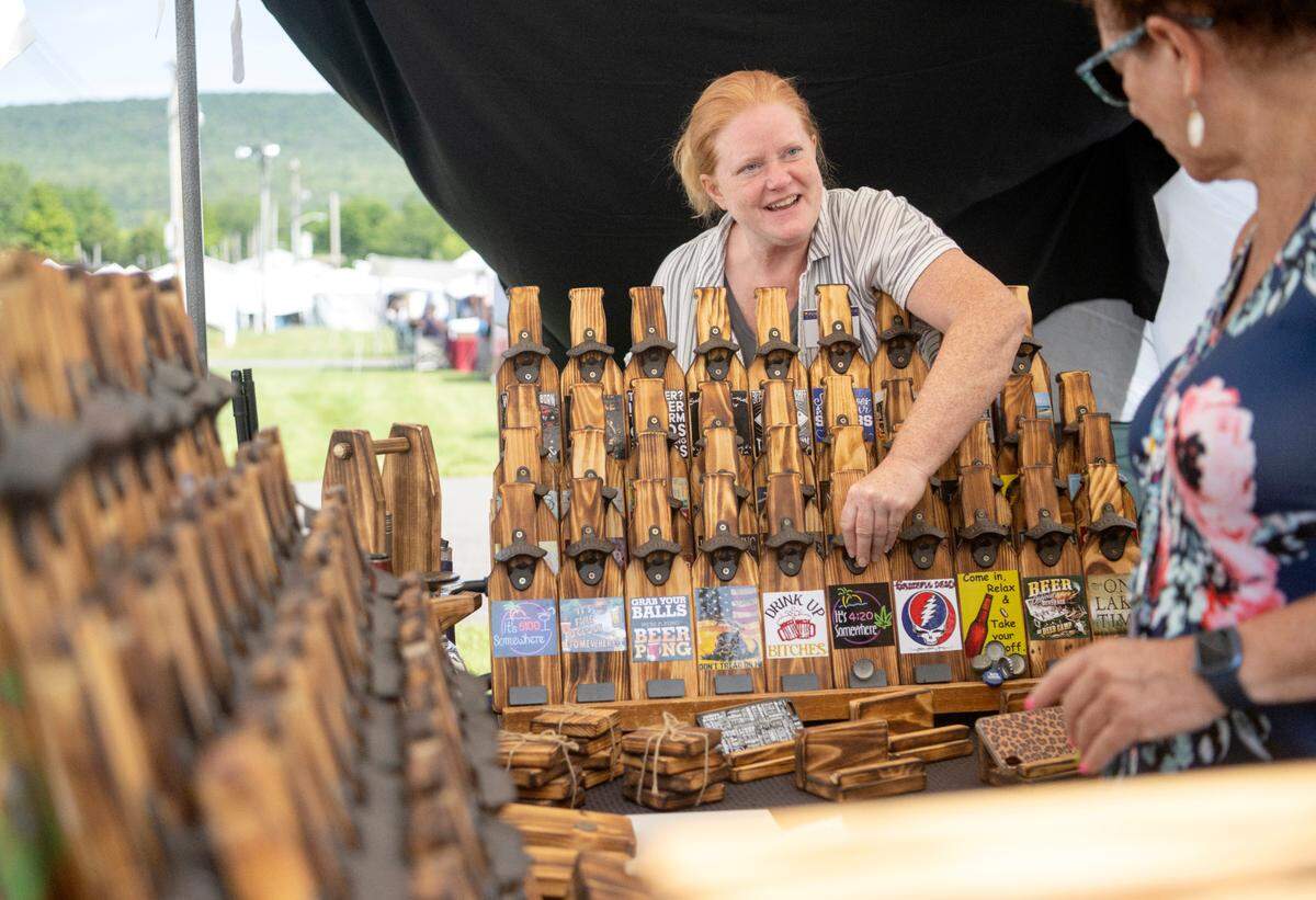 Bonni Oliver talks about her wooden bottle cap openers and games that she makes out of pine as a customer browses her booth at the People’s Choice Festival on Friday, July 14, 2023.