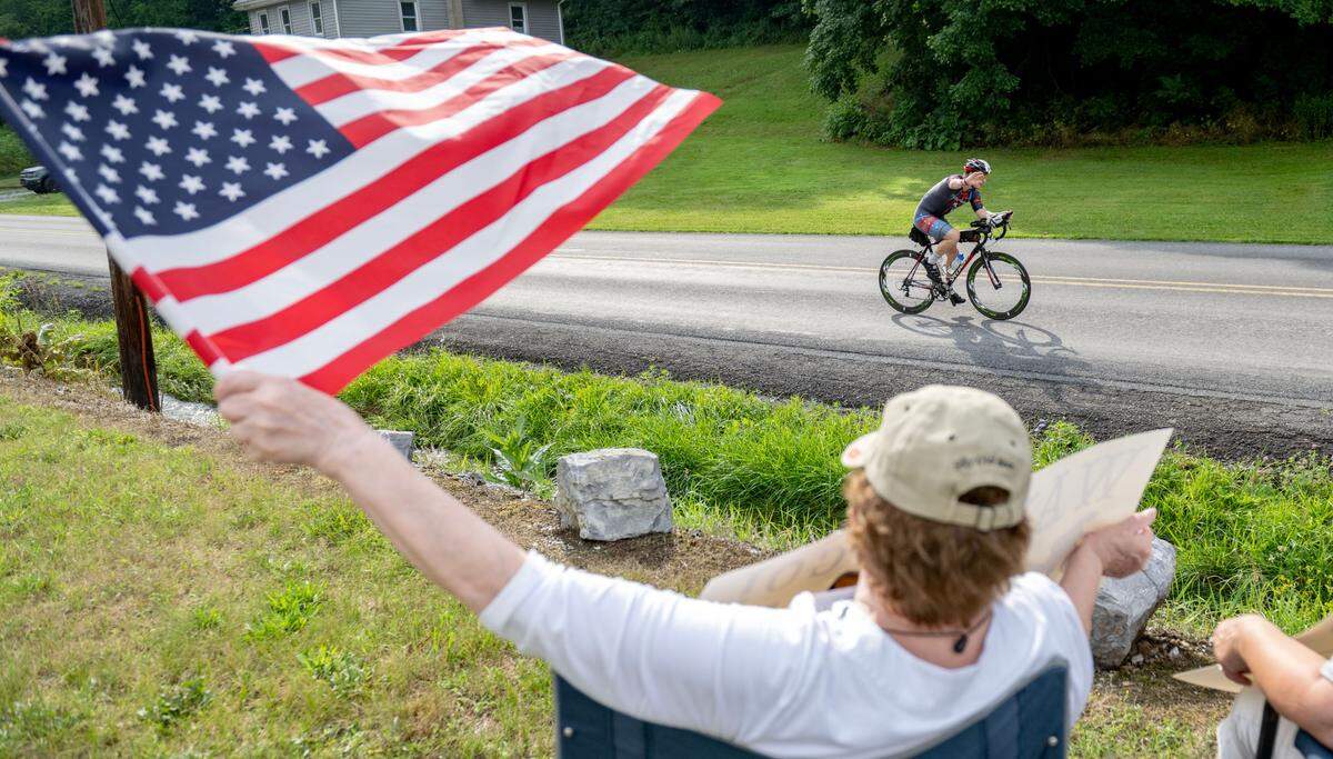 Hope Kmisely waves an American flag and cheers for cyclists they ride along Jacksonville Road in the Ironman 70.3 Pennsylvania Happy Valley on Sunday, June 30, 2024.
