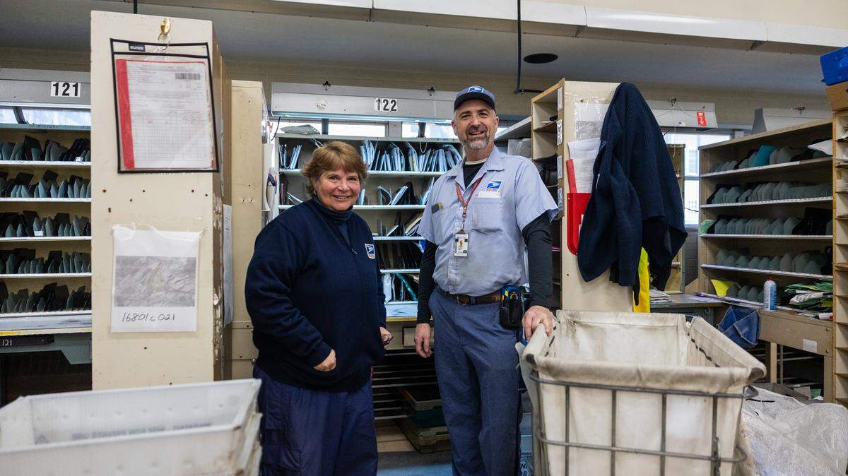 U.S. Postal Service mail carriers Colleen Sweeney, left, and Michael Frank, right, are seen in front of their prep stations inside the State College post office before they start their deliveries on Jan. 13, 2026 in State College, Pa. Sweeney and Frank have been recognized for traveling over around a million miles while working as USPS mail carriers over the course of just over 30 years.