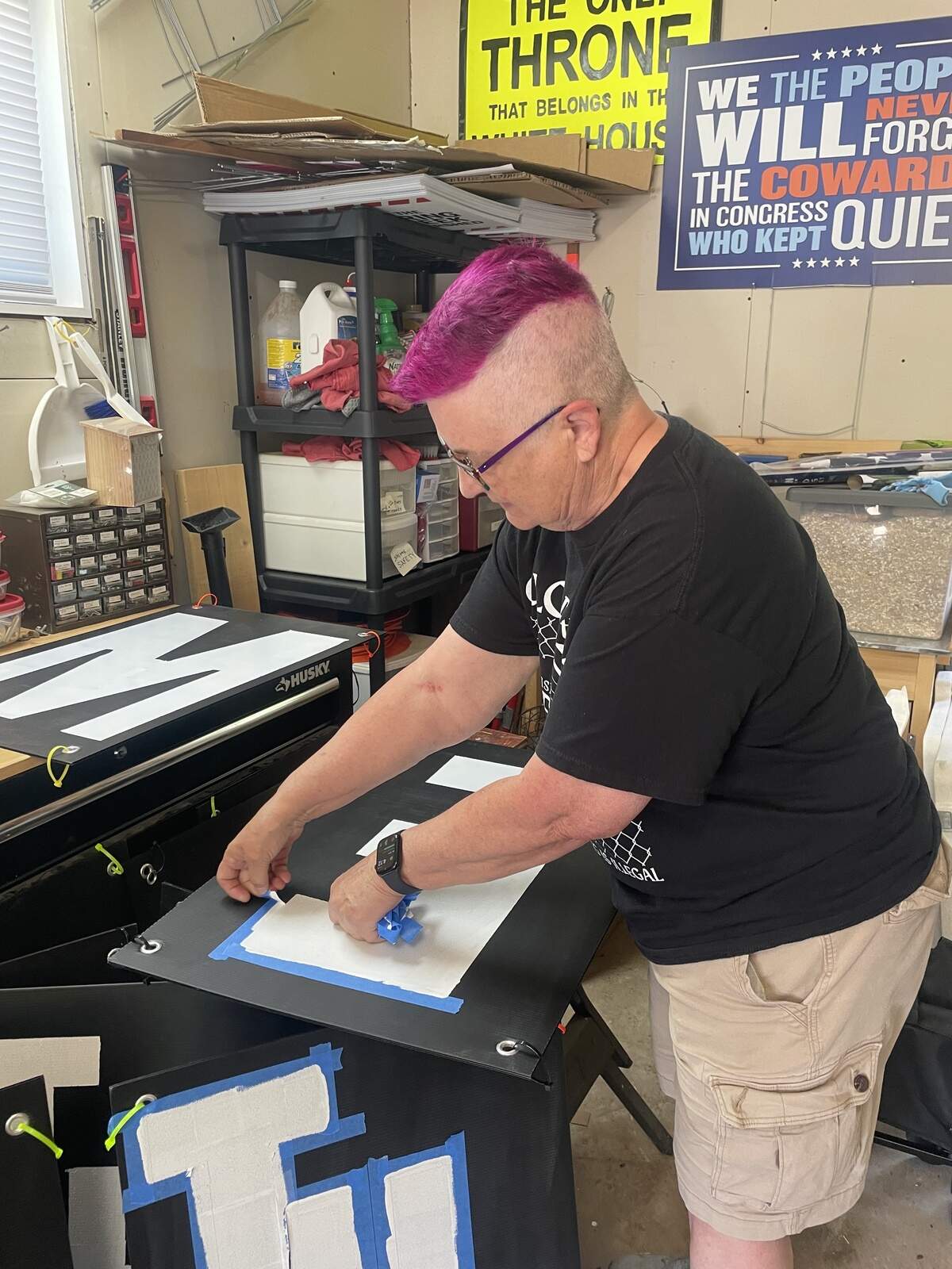 woman removes blue masking tape from black sign with white letter E in garage