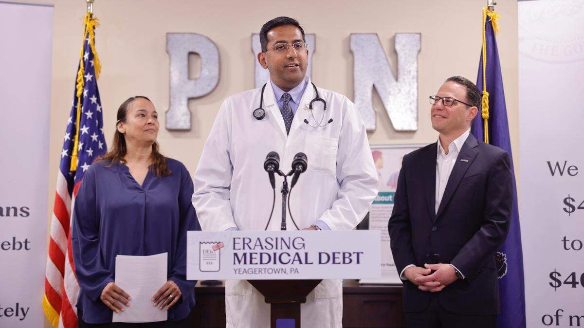 State Rep. Arvind Venkat, flanked by Gov. Josh Shapiro (right) and a resident affected by medical debt (left) stands at a podium for a 2024 event announcing a medical debt relief proposal.
