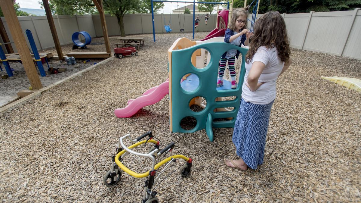 Lucy Reed, 5, stands on the playground with teacher Kaitlyn Mejia as her classmates swing on Tuesday at Easterseals.  Lucy, who uses a walker due to her spina bifida, has a hard time getting around on the mulch playground.
