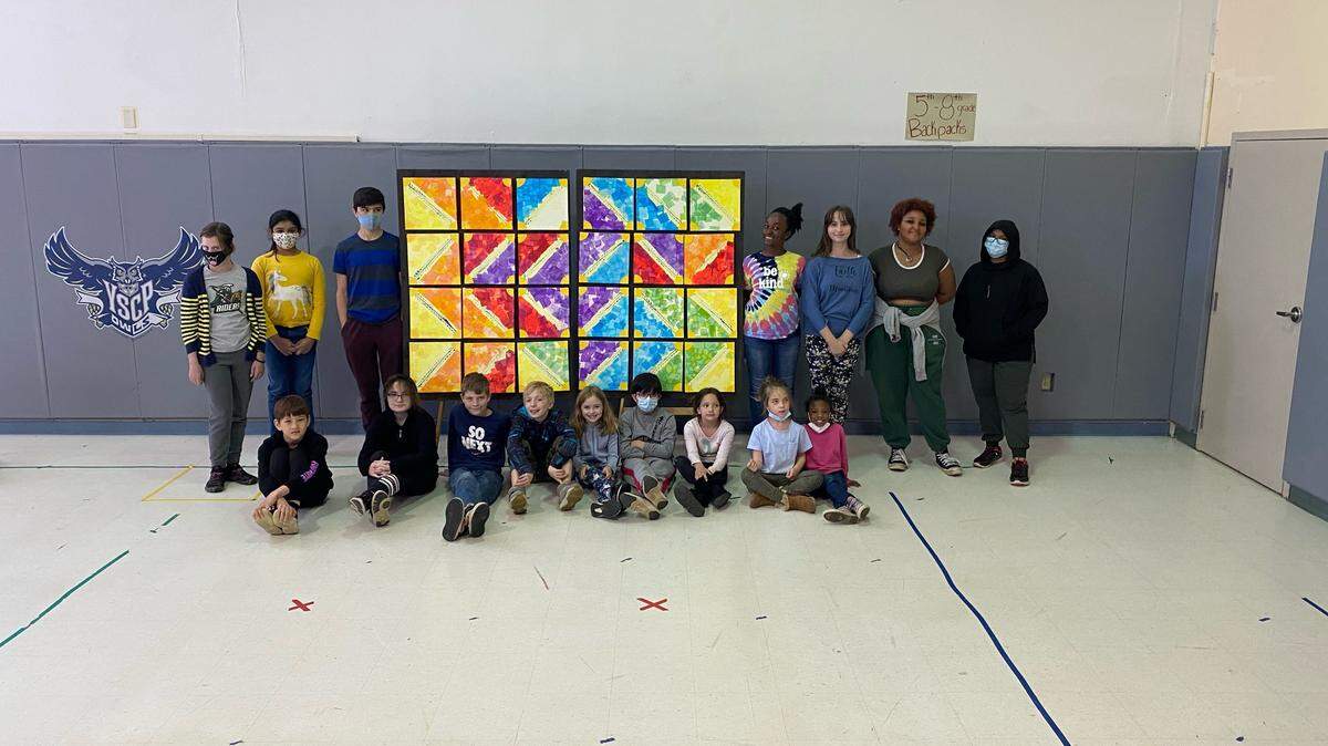 Twenty four students from kindergarten through eighth grade stand in front of the canvases they made as part of a permanent art installation at Young Scholars.