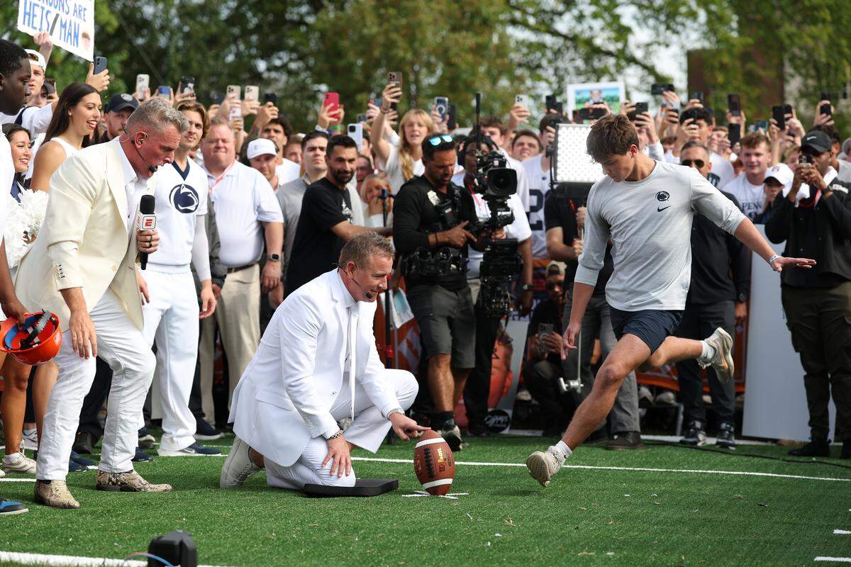 College GameDay analyst Kirk Herbstreit holds the ball for Penn State student Carson Albright, while Pat McAfee looks on, Saturday morning during the Field Goal Kicking Challenge segment on the set of College GameDay at Penn State.