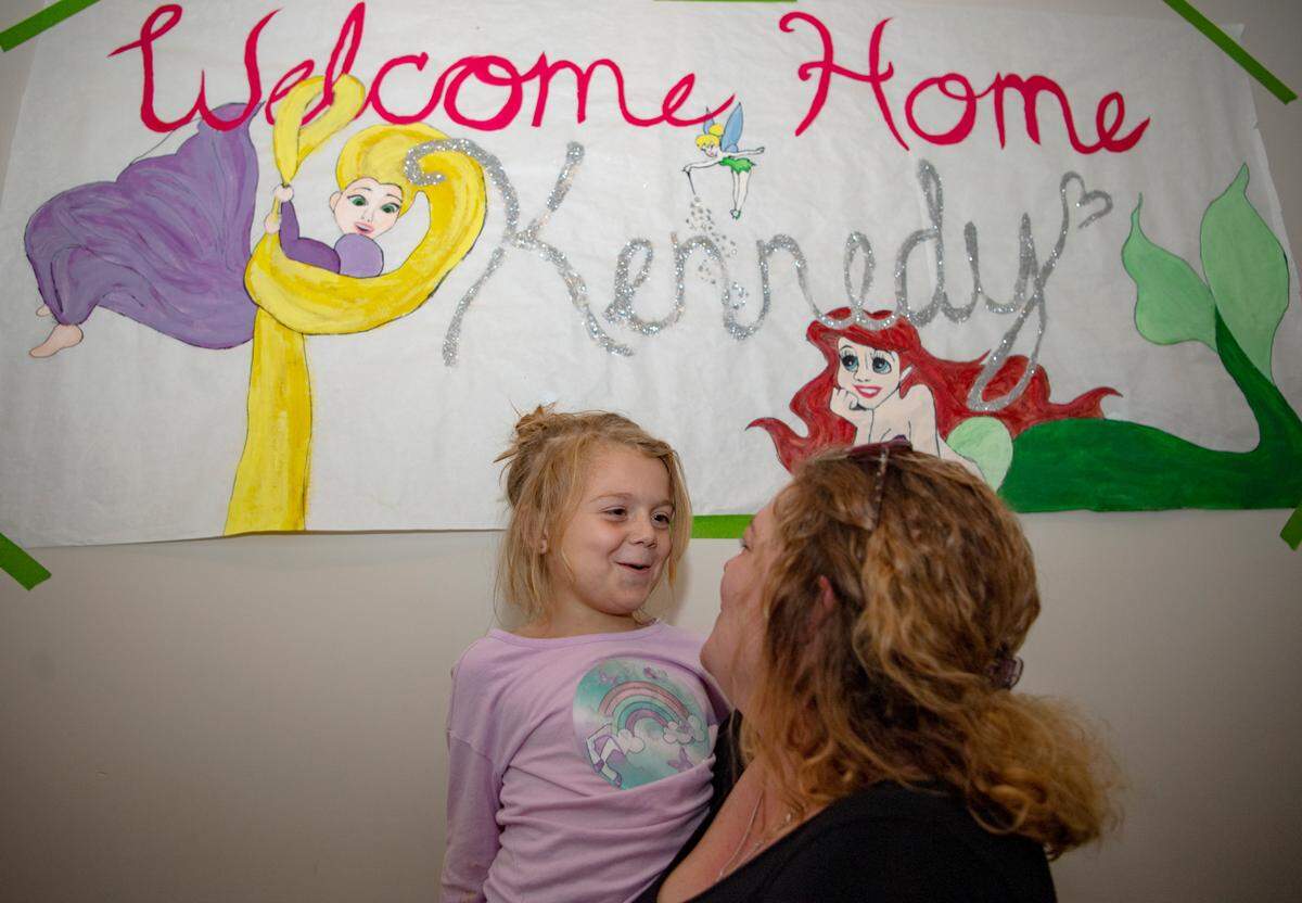 Kennedy Wilson tells her mom a joke next to the banner that welcomed her home from the hospital in 2019. It still hangs in the hallway of the family’s Bellefonte home.