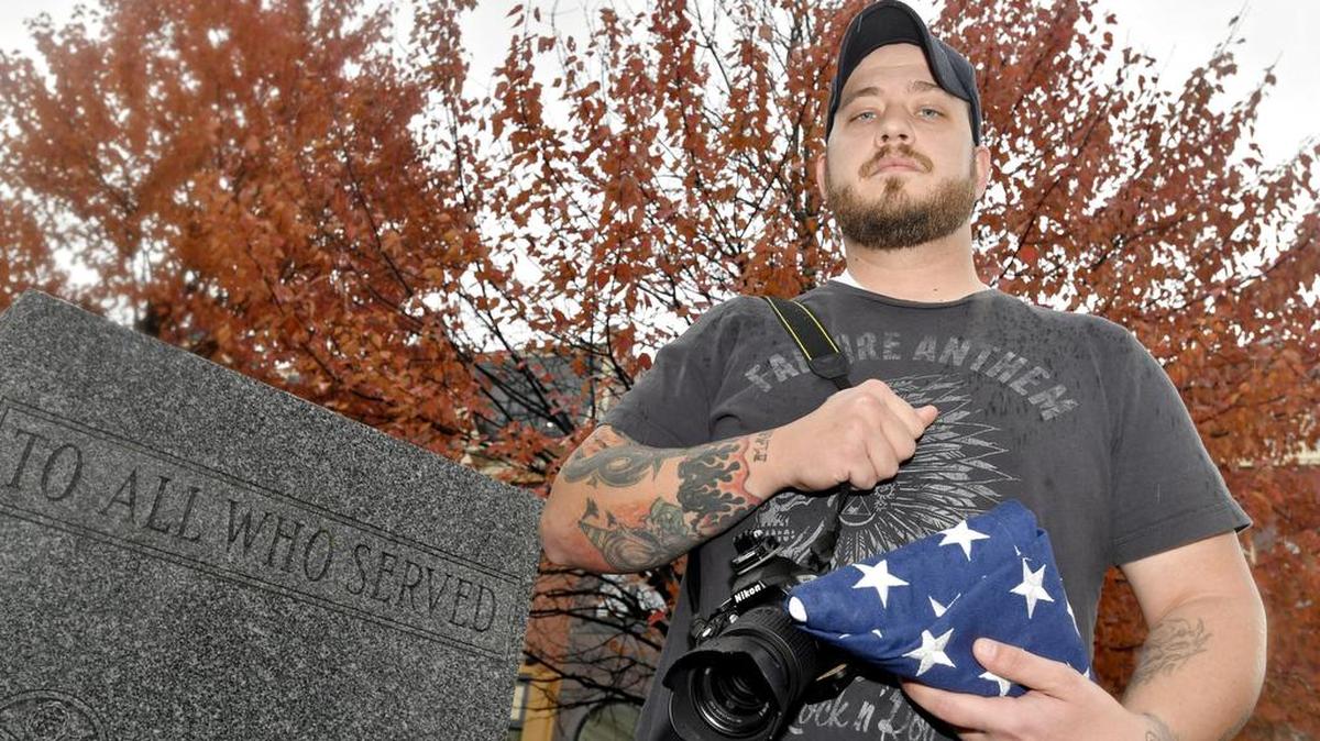 Brian Knepp, a veteran and concert photographer, poses at the veterans memorial in Bellefonte on Friday with his camera and an American flag he was given.