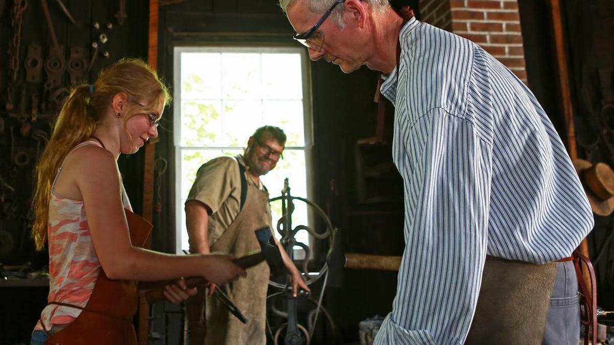 John Wood, of Wilson, N.C., right, helps Mount Nittany Middle School fifth-grader Maeve Flanagan form the end of a fork as Eric Johnson stokes the furnace in the Boalsburg blacksmith shop during Boalsburg Memorial Day festivities on Monday.